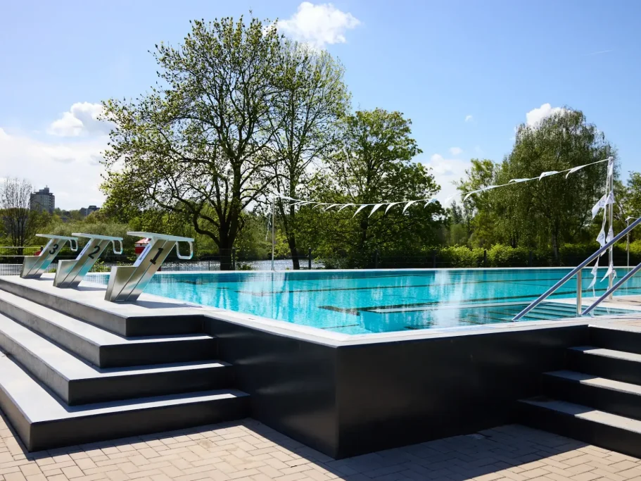 Schwimmbecken mit Treppen und Bäumen im Hintergrund, unter blauem Himmel.