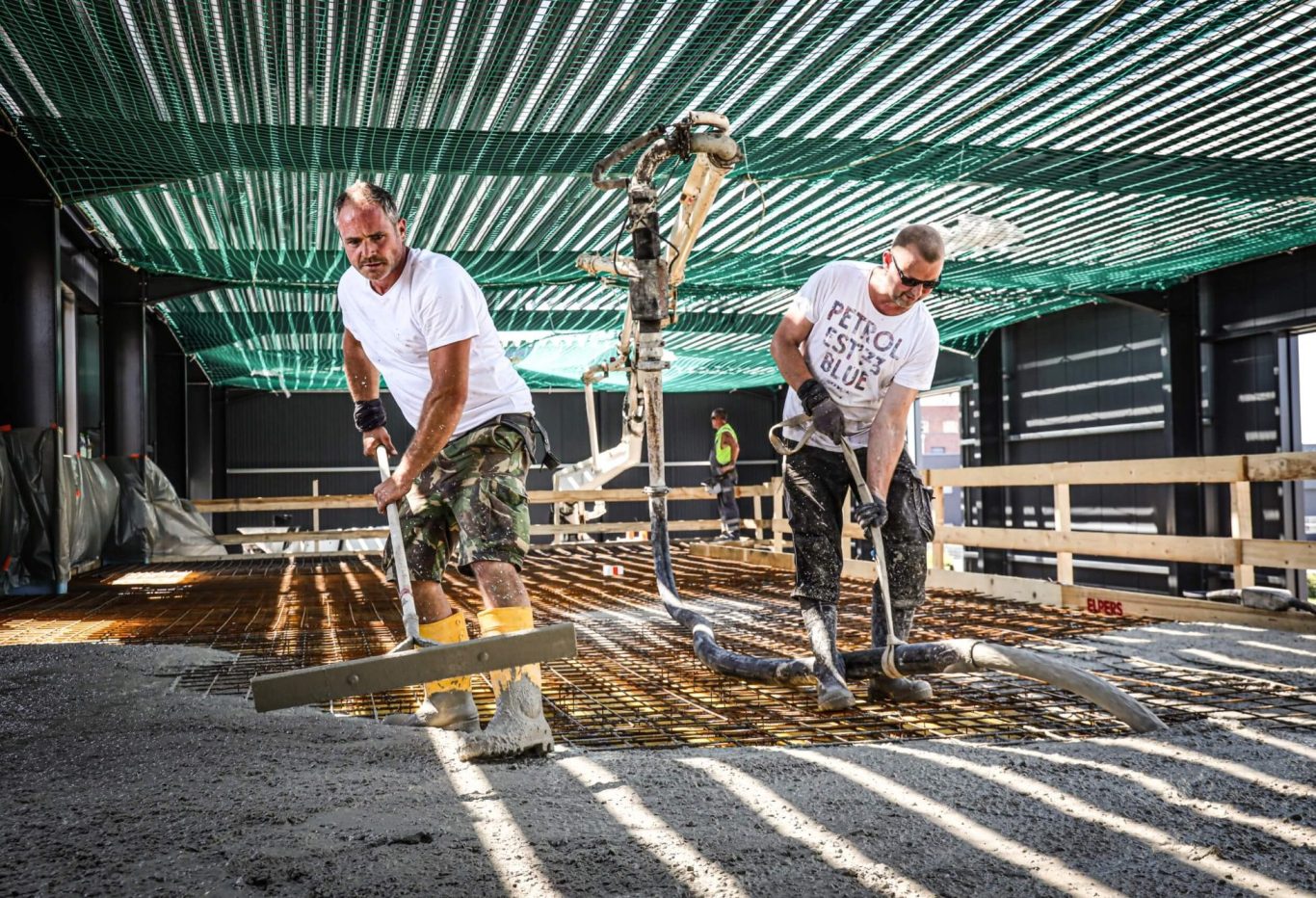 Zwei Bauarbeiter verlegen Beton auf einer Baustelle unter einem grünen Dach.