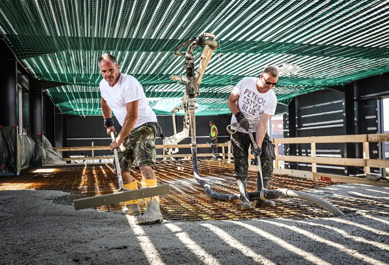 Bauunternehmen Elpers Bau Zwei Bauarbeiter verlegen Beton auf einer Baustelle unter einem grünen Dach.