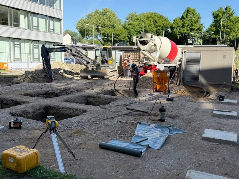Bauarbeiten mit Baustellenfahrzeugen und Betonmischer, Gruben im Boden sichtbar.